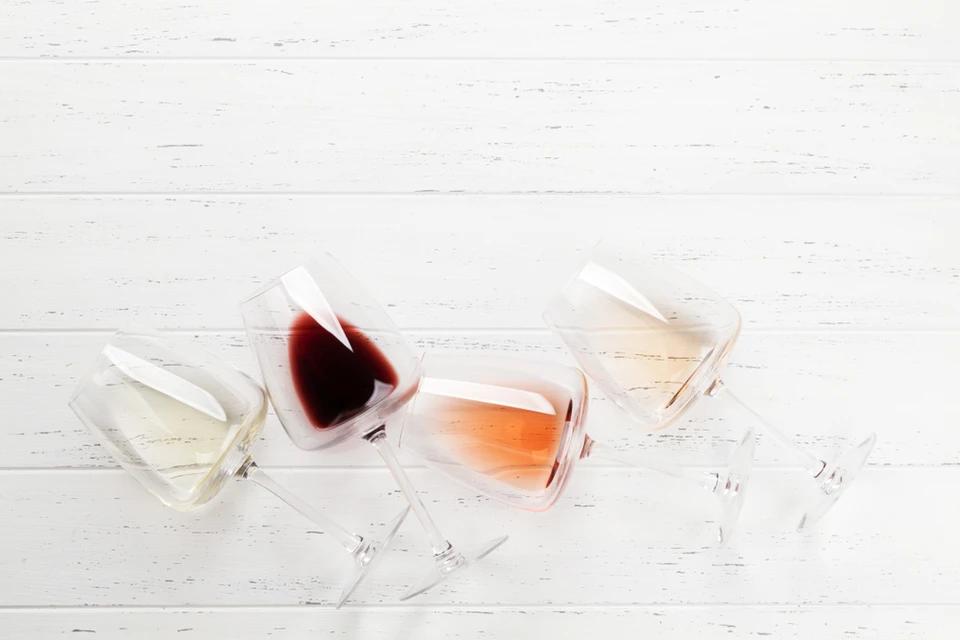 Quatre verres à vin renversés sur surface en bois blanc patiné, contenant différents types de vins : blanc, rouge foncé, rosé et champagne